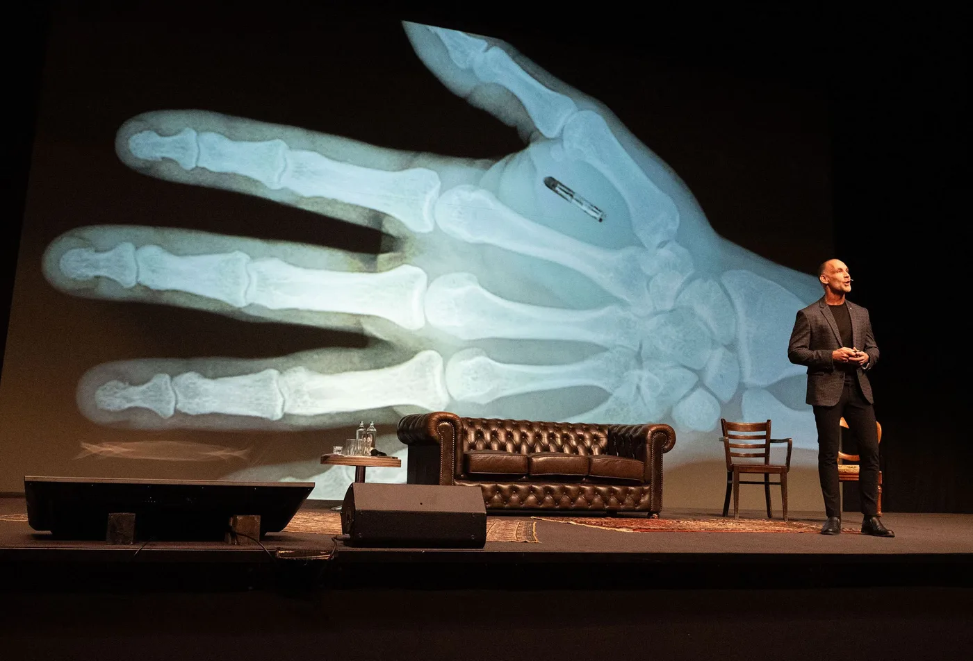 Richard van Hooijdonk op een theaterpodium met chesterfieldbank, naast een grote röntgenfoto van een hand met een chip