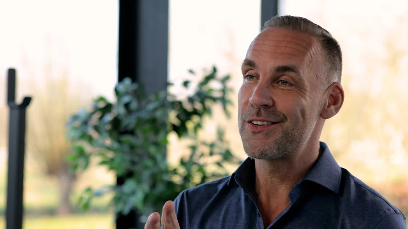 Richard van Hooijdonk in donkerblauw overhemd, pratend tijdens een interview, met een plant en ramen op de achtergrond