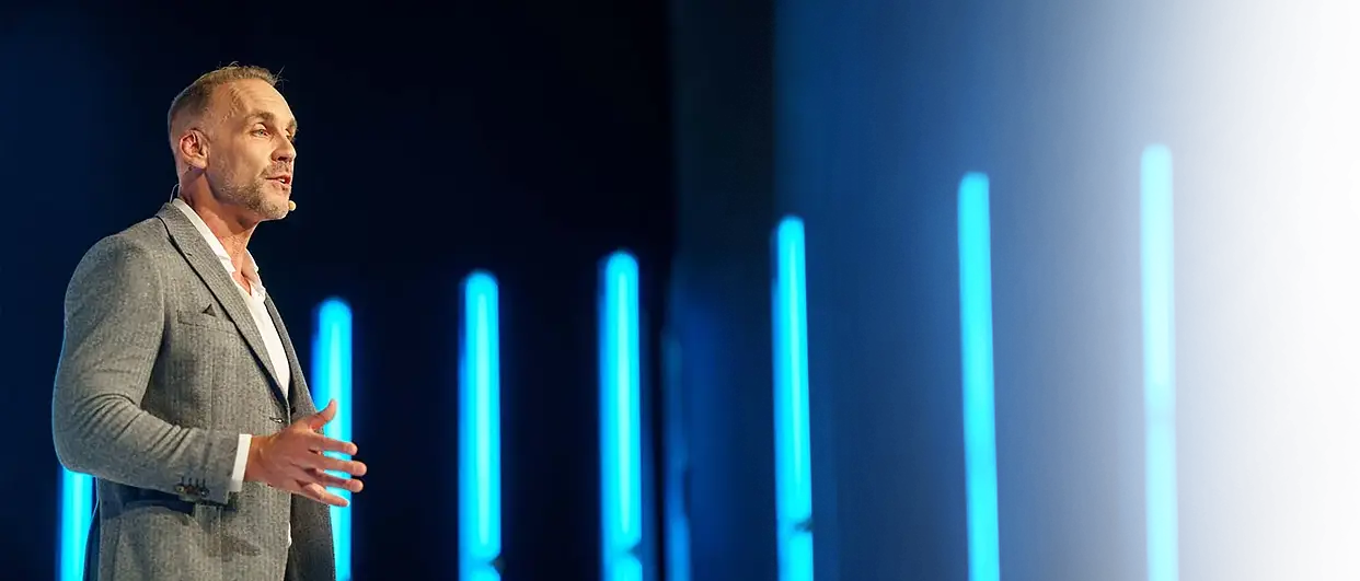 Richard van Hooijdonk in a grey jacket on stage, in front of vertical blue light bars
